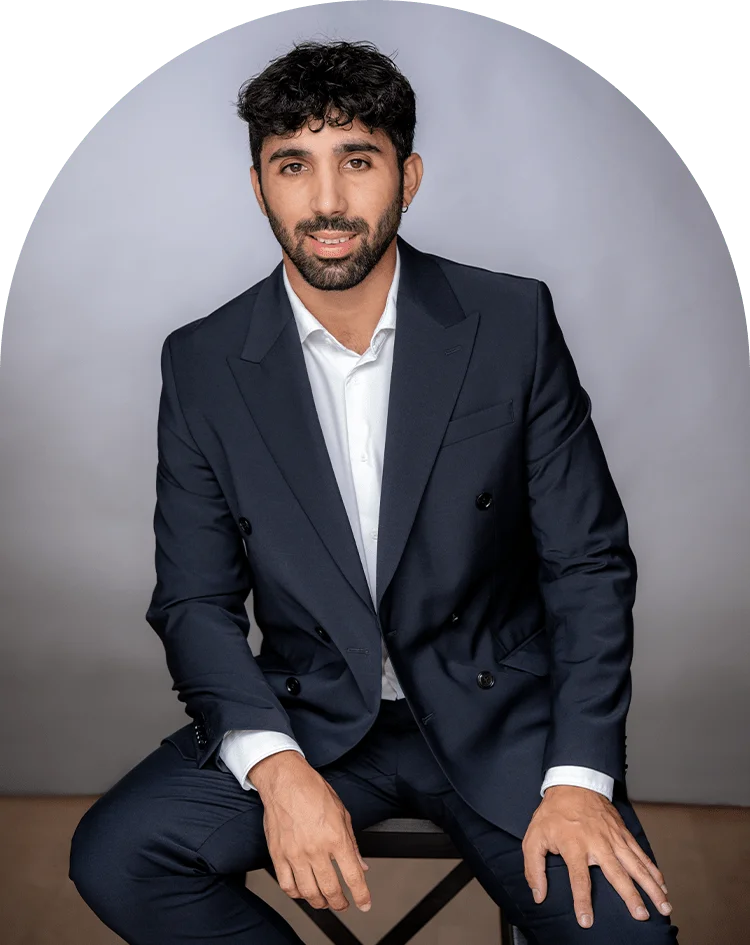 Un homme aux cheveux bruns courts et à la barbe, vêtu d'un costume bleu marine et d'une chemise blanche, est assis sur un tabouret sur un fond gris clair, faisant face à l'appareil photo avec un léger sourire - ce qui donne l'air professionnel d'un expert-comptable ou d'un dentiste.